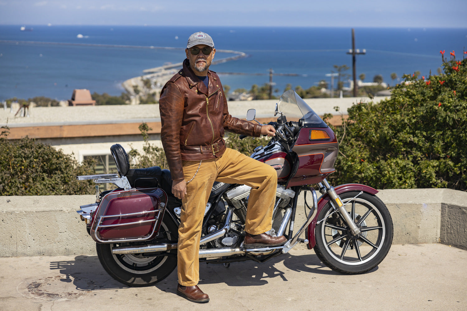 TT&CO. CEO posing next to a vintage-style motorcycle with an ocean view in the background. He is wearing a brown leather jacket, mustard-colored pants, brown boots, sunglasses, and a gray cap.